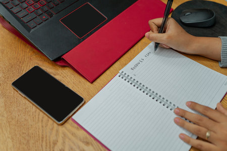 A hand diligently writing detailed notes in a notebook positioned beside a laptop and smartphone