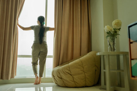 A woman is savoring a delightful and peaceful morning by opening the curtains in her cozy homeの写真素材