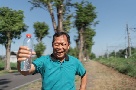 A joyful man showcases a bright water bottle on a sunny day along a rural pathway