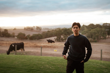 A young man stands in a serene rural setting with cows grazing nearby.