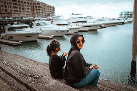 Mother and daughter relax at the marina, surrounded by yachts and calm waters.の写真素材