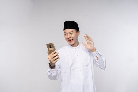 Man in traditional outfit waving at phone during video call, holding gold device and smiling warmly,...