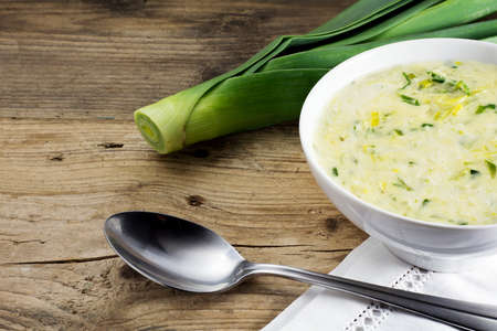 vegetable cream soup in a white ceramic bowl, spoon and fresh leek on rustic woodの写真素材