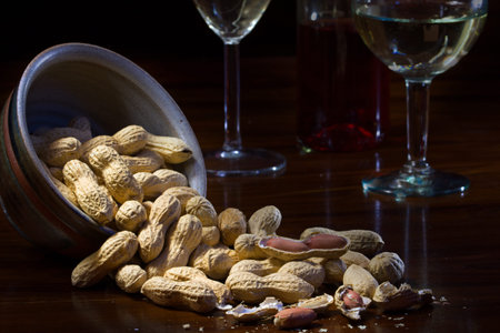 peanuts in a ceramic bowl and on a dark wooden table, wine glasses in the background, evening scene...