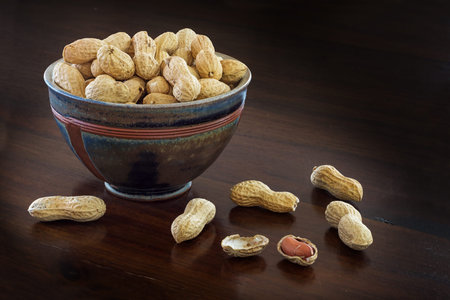 peanuts in shells  in a handmade ceramic bowl on a  brown table, copy space in the dark background