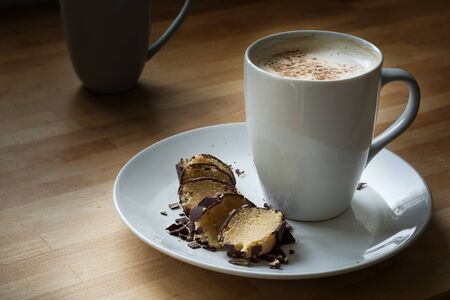 coffee mug with milk foam and cocoa on a white plate with marzipan coated in dark bittersweet chocolateの写真素材