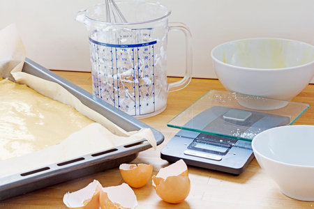 baking utensils, baking tray with dough, plastic jug, kitchen scales, bowls and eggshells on a...