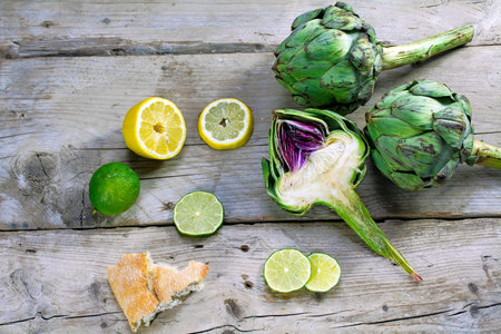 artichokes whole and halv, showing the heart, with lemons, limes and bread on an old weathered wooden tableの写真素材