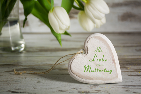 heart shape of wood in front of white tulips on a gray  rustic wooden table, german text Alles Liebe zum Muttertag, that means All Love to your Mothers Day, closeup with selected focus and narrow depth of fieldの写真素材