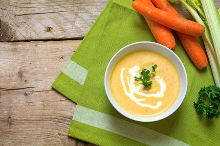 carrot soup with cream and parsley garnish in a bowl on rustic wood, view from above, green napkin, raw carrots and celery in the background, copy spaceの写真素材