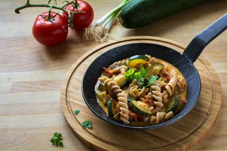 Vegetables with whole grain pasta in a rustic pan on a wooden kitchen board, healthy vegetarian meal to lose weight with zucchini; tomatoes and spring onions, copy space, selected focus, narrow depth of fieldの写真素材