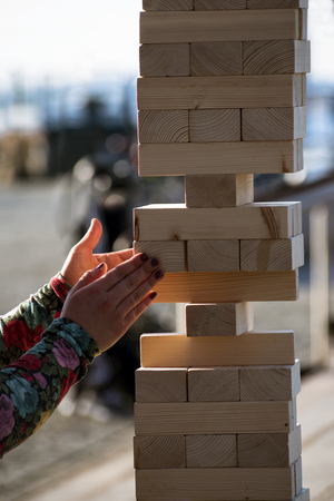 Jenga, girl's hands try to pull out a wooden block, without tipping the tower, group game of physical skill with big blocks for outdoors, verticalの写真素材