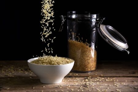 rice grains falling into a white bowl beside a glass jar with rice on a rustic wooden table against a dark background, food concept against hunger and for world nutrition, selected focusの写真素材