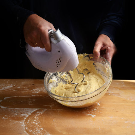 Female hands stirring dough with a mixer for cake or bread in a bowl on a wooden kitchen worktop,...