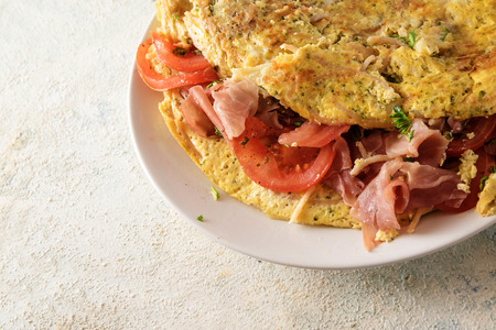 vegetable omelette filled with tomatoes, prosciutto ham and parsley garnish on a plate, light gray background with copy spae, close upの写真素材