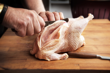 Men's hands prepare a raw chicken with a kitchen knife for cooking on a rustic wooden butcher block, selected focus, narrow depth of fieldの写真素材