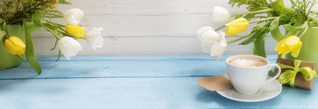 coffee cup, yellow and white tulip flowers in green vases and a little gift on a blue wooden table,...