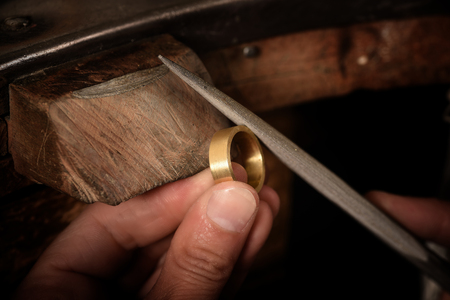Goldsmith hand holds a golden ring on the wooden workbench and works on it with a metal file, close up with copy space,  focus, narrow depth of fieldの写真素材