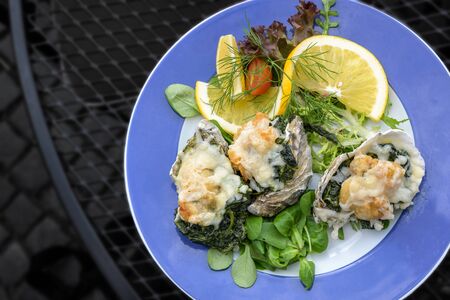 Baked oysters Rockefeller au gratin with parmesan cheese and spinach, served with salad on a blue plate on a dark metal table, high angle view from above, copy space, selected focus, narrow depth of fieldの写真素材