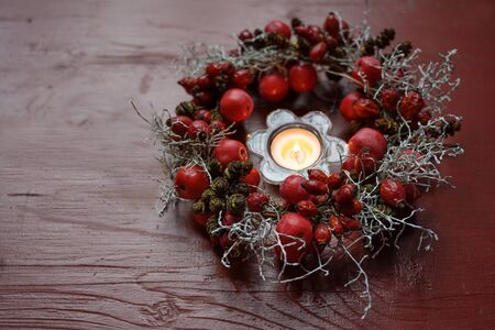 Natural wreath with rose hips and small apples around a burning candle on a red wooden tableの写真素材