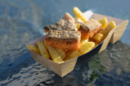 fish and chips with fried smoked salmon and french fries in a cardboard box on a glass table