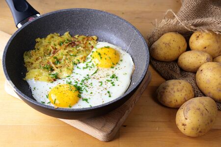 Fried eggs and Swiss roesti in a pan on a wooden kitchen table with a sack of potatoesの写真素材