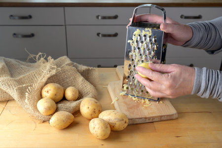 Hands of a woman are grating potatoes with a metal grater on a wooden kitchen table, selected focus,...