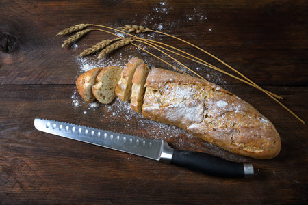Baguette or French bread half sliced, some wheat ears and a knife on a dark rustic wood
