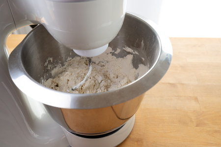 Electric stand mixer is stirring raw dough with the dough hook in a stainless steel bowl on a wooden...