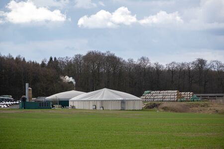 Biogas plant with straw bale stock and smoking chimney on the field at the edge of the forest, cloudy sky, copy spaceの写真素材