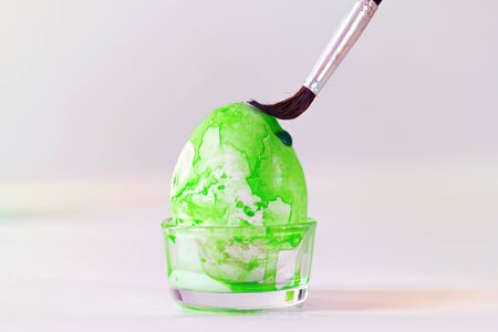 Brush is painting green watercolor on an Easter egg for the holiday decoration, light grey background with copy space, selected focus, narrow depth of fieldの写真素材