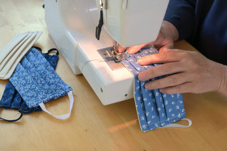Hands of an older woman are sewing a community face mask during the coronavirus pandemic, homemade...