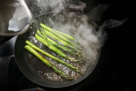 Green asparagus in a frying pan is poured with water, that produces a lot of steam, black stove with copy space, selected focus, narrow depth of fieldの写真素材