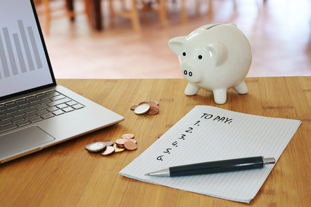 Calculating household costs, laptop, coins, a piggy bank and a list what to pay on a table in the...