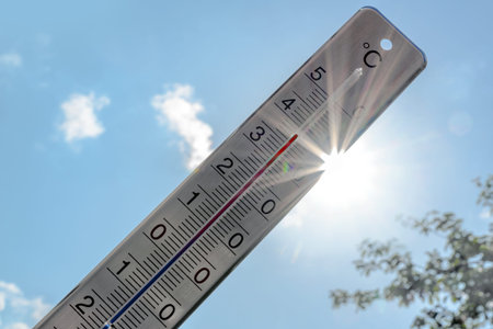 Thermometer shows heat in the summer season against a blue sky with sunbeams and lens flares,...