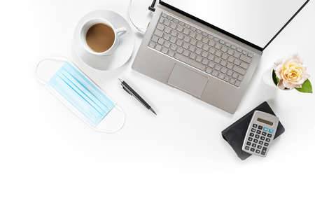 White office desk with laptop, working utilities, coffee cup and a surgical face mask against coronavirus and covid-19 infection, new normal business concept, copy space, high angle view from aboveの写真素材