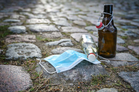Beer and liquor bottles and a surgical face mask left on a cobblestone road, while bars have closed due to infection with coronavirus and covid-19, some party people drinking in the streets, copy space, selected focus, very narrow depth of fieldの写真素材