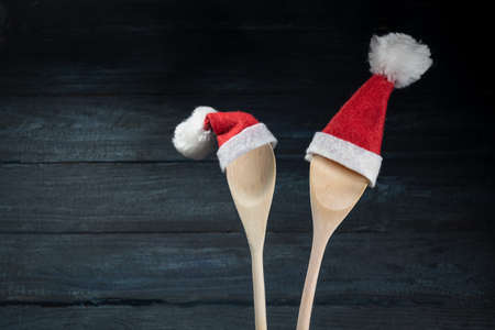 Two cooking spoons with red Santa Claus hats against a dark blue wooden background, Christmas invitation for a dinner or greeting card with copy space, selected focusの写真素材