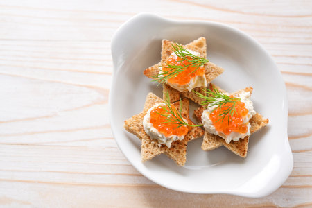 Canapes with red caviar on toasted bread in star shape with cream and dill garnish on a white plate, light wooden table, copy space, high angle view from above, selected focusの写真素材