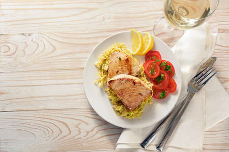 Fried tuna steaks on savoy cabbage, tomatoes and lemon served with wine on a plate and a white painted wooden table with napkin and cutlery, copy space, high angle view from directly above, selected focusの写真素材