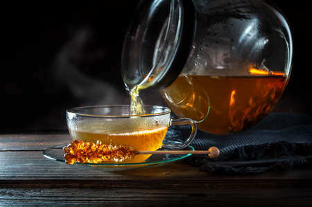 Hot steaming tea is poured from a pot into a glass cup, served with a stick of rock candy, motion blur, dark rustic wooden background with copy space, selected focus, narrow depth of fieldの写真素材