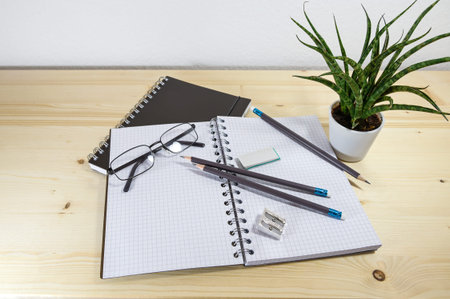 Office or study desk with an open spiral notebook, pencils, glasses and supplies on a wooden table,...