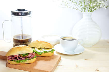 Sandwich rolls with salami and eggs, fresh hot coffee and a flower vase on a wooden table for breakfast or lunch break, light background with copy space, selected focus, narrow depth of fieldの写真素材