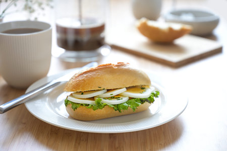 Sandwich roll with egg slices, lettuce and chives garnish on a white plate and fresh coffee on a...