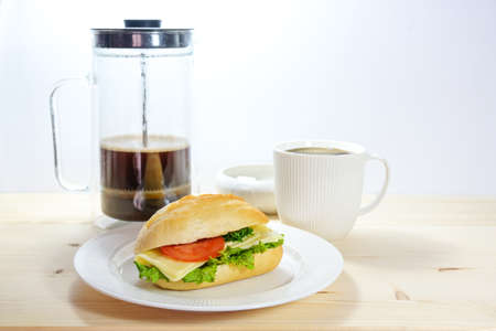 Sandwich bun with cheese, lettuce and tomato on a plate, coffee pot and cup, wooden table and light background, copy space, selected focus, narrow depth of fieldの写真素材