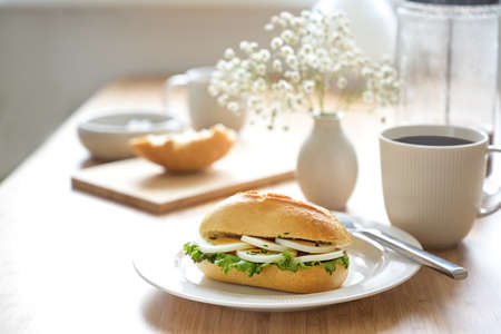 Breakfast table set with egg slices in a sandwich roll, coffee cup and a small flower bouquet, light background with copy space, selected focus, narrow depth of fieldの写真素材