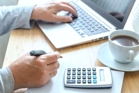 Hands of an elderly woman working on a laptop and a calculator for business accounting on a wooden desk with a coffee cup, copy space, selected focus, narrow depth of fieldの写真素材