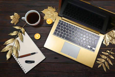 golden laptop computer, gold painted autumn leaves, coffee cup and a notepad with pen on a dark rustic wooden table, holiday decoration on the home office desk, flat lay from above, copy space, selected focusの写真素材