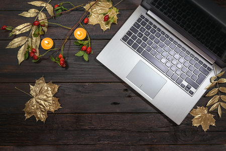 silver laptop computer with gold painted autumn leaves, rose hips and candles on a dark rustic wooden table, seasonal business in the home office, flat lay from above, copy space, selected focusの写真素材