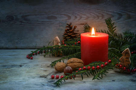 Warm light on a cold winter night from a red candle with flame, evergreen yew branches, nuts and Christmas decoration on blue gray rustic wooden planks, copy space, selected focus, narrow depth of fieldの写真素材